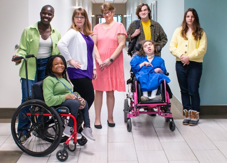 Four women and one man standing in a hallway with two women seated in wheelchairs, all looking towards the camera and smiling.