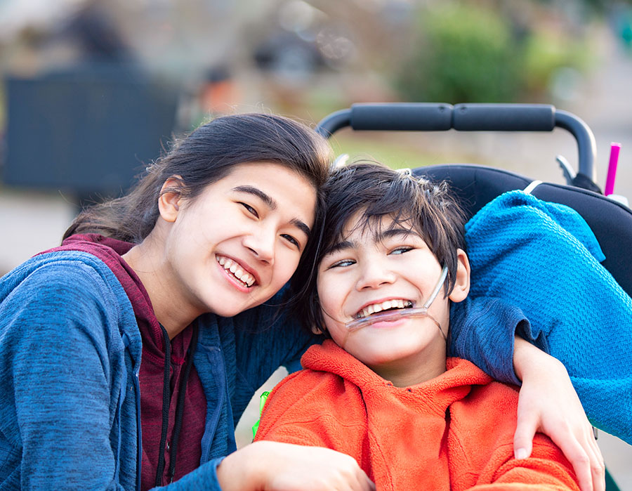 Big sister hugging disabled brother in wheelchair outdoors, smiling